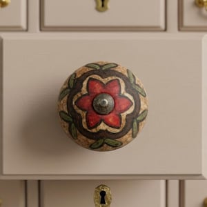 May include: A decorative cabinet knob with a floral design. The knob is round with a tan background, a red flower, green leaves, and a silver center. The knob is attached to a light beige cabinet door.