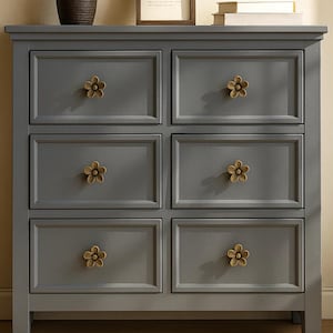 May include: A gray six-drawer dresser with flower-shaped brass knobs. The dresser has a simple, classic design, with each drawer featuring a single knob. The knobs are a warm brass color, contrasting with the cool gray of the dresser.
