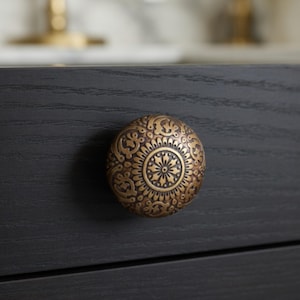 May include: A round, antique brass cabinet knob with an ornate floral design. The knob is mounted on a dark gray wooden drawer, showcasing its intricate details and warm metallic finish. This decorative hardware adds a touch of vintage charm.