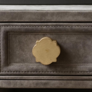 May include: Close-up of a gray, upholstered drawer with a gold-colored, ornate knob. The drawer has a rectangular recessed panel and is stitched with a contrasting thread. The knob is a stylized quatrefoil shape.