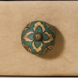 May include: A round, teal and beige decorative cabinet knob with a floral design. The knob has a bronze-colored center and is set against a tan background. The knob is likely made of ceramic or wood.