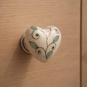 May include: Heart-shaped cabinet knob with a cream-colored base and a hand-painted design of green leaves and black vines. The knob is mounted on a light-colored wooden surface, offering a decorative touch for furniture.