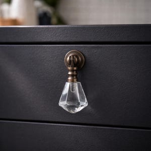 May include: A close-up of a black drawer with a crystal-shaped knob. The knob has a bronze-colored base and a clear, faceted crystal handle. The drawer has a textured surface.