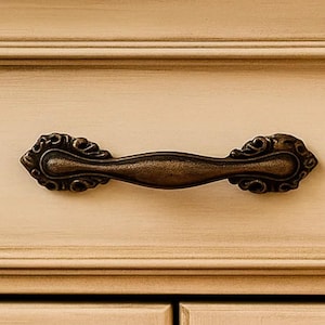 May include: Bronze-toned cabinet pull with ornate, scroll-like detailing on the ends. The handle is a curved bar shape, and the hardware is mounted on a light beige cabinet door. This decorative hardware adds a vintage touch.