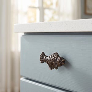 May include: A dark bronze-toned metal cabinet knob shaped like a fish with detailed scales, fins, and a defined eye. The hardware is mounted onto a light blue painted wooden drawer front, featuring a horizontal grain and a smooth, matte finish.