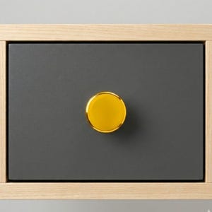 May include: A close-up of a drawer with a dark gray front and a gold-colored, round knob. The drawer is set within a light-colored wooden frame. The knob is centered on the drawer front.