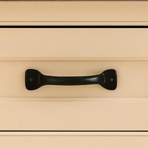 May include: A black, horizontal drawer pull with a curved handle and rounded ends. The pull is mounted on a cream-colored cabinet drawer with horizontal paneling. The hardware is centered and appears to be made of metal.