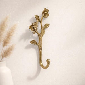 May include: A decorative brass wall hook featuring a floral and bee design. The hook is shaped like a branch with leaves, a bee, and a flower. The hook is mounted on a white wall, adding a touch of nature-inspired decor.