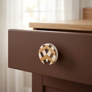 May include: A circular cabinet knob featuring a geometric chevron pattern composed of alternating light and dark wood segments inlaid within a smooth white resin base, mounted on the front of a solid dark brown wooden drawer front.