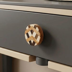 May include: Close-up of a gray drawer with a decorative wooden knob. The knob is round with a chevron pattern in shades of brown, beige, and dark wood. The drawer is part of a larger piece of furniture with beige trim.
