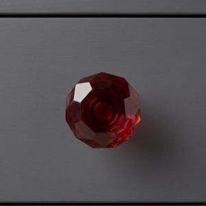 May include: A faceted, ruby-red cabinet knob. The knob has a geometric design with multiple flat surfaces that reflect light. The knob is mounted on a gray surface, creating a contrast between the vibrant red and the muted background.