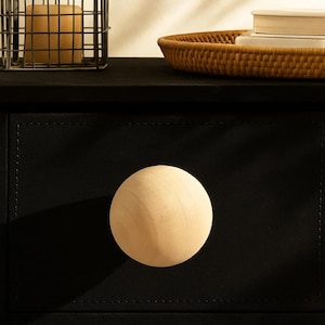 May include: Close-up of a black drawer with a large, round, light-colored wooden knob. The drawer is part of a larger black piece of furniture. A woven tray and a wire basket are visible in the background.
