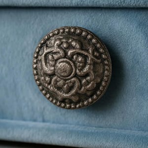 May include: Close-up of a decorative, antique-style metal knob with an ornate floral design. The knob is circular with a detailed pattern and a dark, aged finish. It is set against a soft, light blue fabric background.