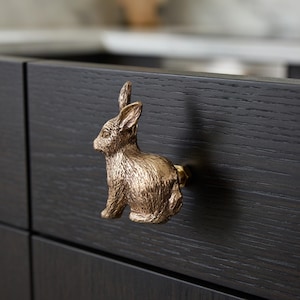 May include: A bronze-colored bunny-shaped cabinet knob. The knob is attached to a dark brown cabinet door. The bunny is sitting upright with its ears perked up. The knob adds a decorative touch to the cabinet.