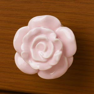 May include: A light pink ceramic rose-shaped cabinet knob. The knob has a glossy finish and is set against a brown wooden surface. The rose design features multiple petals and a detailed center.