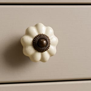 May include: Cream-colored, flower-shaped cabinet knob with a dark brown center. The knob is mounted on a light beige surface, suggesting a piece of furniture. The design is reminiscent of a daisy or other simple flower.
