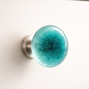 May include: A round, teal glass cabinet knob with a speckled design. The knob is mounted on a brushed nickel base and is set against a white surface. This decorative hardware adds a pop of color.