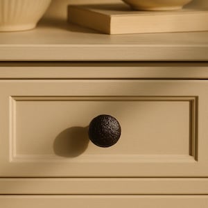 May include: Close-up of a drawer with a textured, dark brown knob. The drawer is painted a light beige color, with a recessed panel design. The knob casts a shadow on the drawer front.