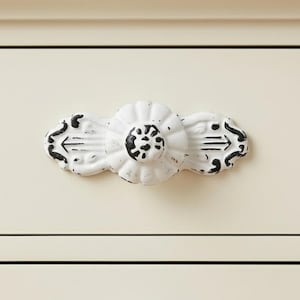 May include: White and black distressed floral cabinet pull. The center of the pull has a flower design, with arrow-like details on each end. The pull is mounted on a cream-colored cabinet.
