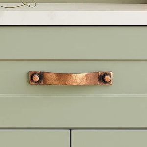May include: Close-up of a copper-colored cabinet pull handle. The handle is a horizontal bar with a slightly curved shape, attached to the cabinet with two round, dark-colored fasteners. The cabinet is painted a light green color.