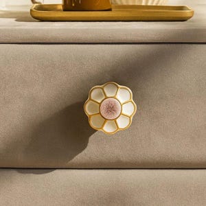 May include: A flower-shaped cabinet knob with a white and gold finish and a pink center. The knob is mounted on a light brown drawer. The knob's design is reminiscent of a daisy.