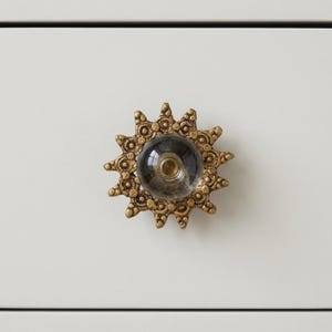 May include: A decorative cabinet knob with a sunburst design. The knob features a clear glass center and a gold-colored metal frame. The frame has an intricate, detailed pattern. The knob is mounted on a white surface.
