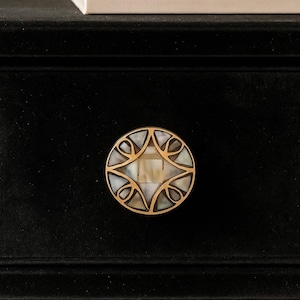 May include: A round, decorative cabinet knob with a gold-colored metal frame. The knob features a central square and four surrounding petal-like shapes, filled with iridescent mother-of-pearl. The knob is set against a black surface.