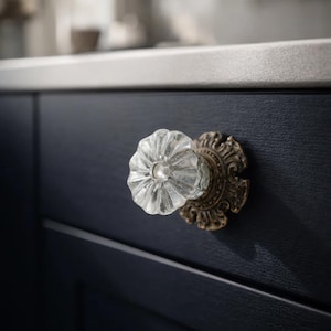 May include: A clear glass cabinet knob with a flower-like design, mounted on an ornate, antique brass base. The knob is attached to a dark blue cabinet door, showcasing a vintage aesthetic.