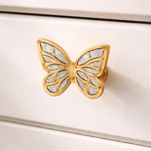 May include: A gold-colored butterfly-shaped cabinet knob with iridescent white accents. The knob is mounted on a white surface. The butterfly design features detailed wing patterns, adding a decorative touch to furniture.