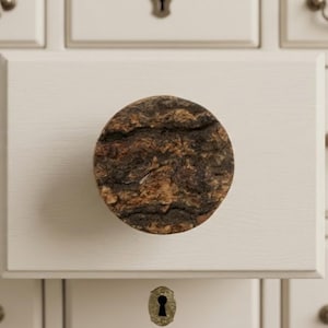 May include: A round, textured cabinet knob made of natural wood, featuring shades of brown and tan. The knob is mounted on a white cabinet door, with a keyhole visible below.