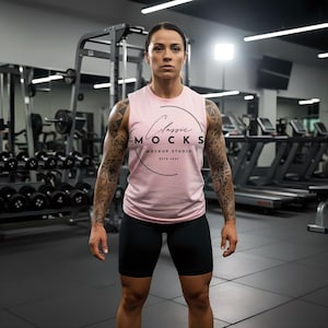 May include: A sleeveless pink tank top with the text "Classic Mocks Mockup Studio" in a black circular design. The person is wearing black shorts and is standing in a gym with weights and exercise equipment.