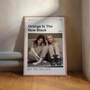 May include: Framed poster featuring the text "Orange Is The New Black" and two women in beige prison uniforms. The poster is in a light wood frame and is displayed against a neutral-toned wall.