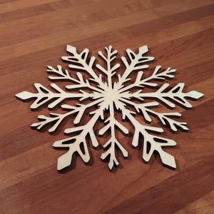 Pode incluir: Um grande floco de neve de madeira, intrincadamente cortado. O floco de neve é de cor clara com um design detalhado, apresentando múltiplas pontas e padrões delicados. É colocado sobre uma superfície de madeira de tons quentes, criando um contraste de cor e textura.