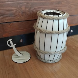 May include: A decorative wooden barrel with vertical black lines and rope accents. The barrel is white and has a cylindrical shape. An anchor-shaped cutout is next to the barrel. The items are on a wooden surface.