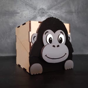 Può includere: Una scatola portaoggetti quadrata in legno con un pannello frontale decorativo a forma di gorilla dei cartoni animati. Il gorilla ha pelliccia nera, un viso grigio chiaro, grandi occhi bianchi e due mani grigio chiaro poggiate sulla base.