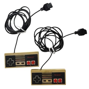 May include: Two rectangular video game controllers with a beige casing and black faceplate. Each controller features a black directional pad, two small rectangular buttons labelled Select and Start, and two round red action buttons on the right side.