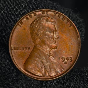 May include: A copper-coloured 1943 Lincoln penny. The coin shows a profile of Abraham Lincoln, with the inscriptions "IN GOD WE TRUST" and "LIBERTY". The year "1943" is visible, along with the mint mark "D".