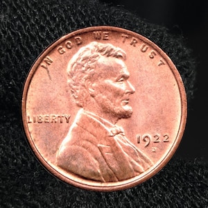 Puede incluir: Una moneda de un centavo Lincoln de 1922 de color cobre. El anverso presenta un perfil de Abraham Lincoln, la palabra "LIBERTY" y la fecha "1922". La moneda también tiene las palabras "IN GOD WE TRUST".