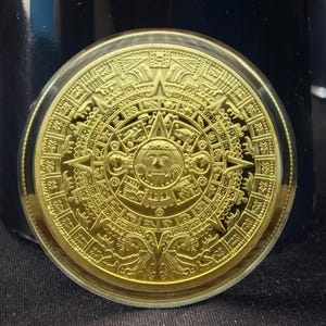 May include: A gold-colored coin featuring a detailed depiction of the Aztec calendar. The coin is round and encased in a clear plastic holder. The design includes intricate patterns and a central sun face.