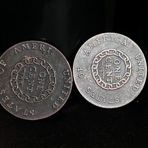 May include: Two antique US one-cent coins. The coins are silver in color and feature the words "AMERICA", "UNITED STATES OF AMERICA", and "ONE CENT". The coins have a chain design around the center.