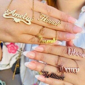 May include: Several gold and rose-gold nameplate necklaces spelling out names such as "Amelia," "Hannah," "Emma," "Olivia," "Remy," and "William." The necklaces are shown on a person's hands.