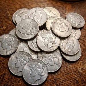 May include: A collection of silver-colored Peace dollars, which are circular coins featuring the profile of Lady Liberty wearing a radiant crown. Each coin is inscribed with the text IN GOD WE TRVST and the year of minting, such as 1921, 1922, 1923, 1925, 1927, and 1928.
