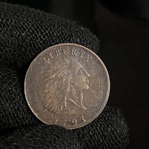 May include: A silver coin with the word "LIBERTY" at the top and the date "1793" at the bottom. The coin features a profile of a woman with flowing hair. The coin is held by a black gloved hand.