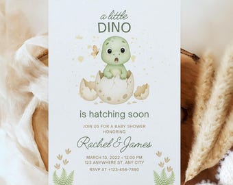 May include: A baby shower invitation featuring a watercolour illustration of a green dinosaur hatching from an egg. The text reads "a little DINO is hatching soon" and includes the names Rachel & James, date, time, and RSVP information. The invitation is surrounded by soft, neutral-toned decor.
