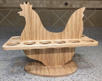 May include: A wooden egg holder in the shape of a hen. The light brown holder has a horizontal shelf with six circular openings for eggs. The hen's body and head are cut out of wood, with a rounded base.