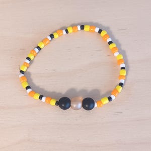 May include: A circular beaded bracelet featuring a repeating pattern of small orange, yellow, white, and black beads. The center of the bracelet is accented with two larger matte black beads flanking a single larger translucent peach-colored bead.