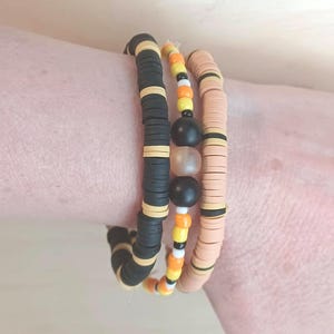 May include: A set of three bracelets featuring flat black and tan clay discs, a central strand of small yellow, orange, white, and black beads with larger matte black and translucent spheres, and a strand of tan discs with black accents.