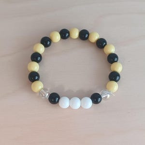 May include: A beaded bracelet featuring alternating black and pale yellow beads. The bracelet also includes three white beads and two clear beads. The bracelet is displayed on a light wooden surface.