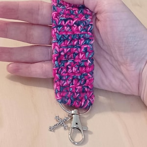 May include: A handmade crochet wrist strap in shades of pink and teal, attached to a silver-colored clasp and keyring. A decorative silver cross charm hangs from the keyring. The strap is designed for holding keys or other small items.