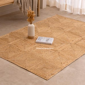 Op de afbeelding: Een rechthoekig jute vloerkleed in natuurlijke tinten, met een geometrisch patroon. Een witte en bruine vaas met gedroogde bloemen en een boek liggen op het vloerkleed. Het vloerkleed ligt op een lichte vloer.